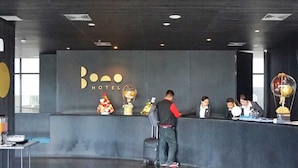 Un espace d’accueil d’hôtel au design moderne, comprenant un comptoir de réception, un grand logo sur le mur et quelques éléments décoratifs.