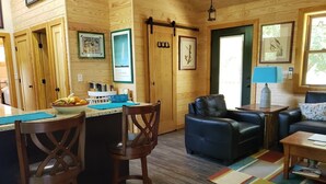 Interior acolhedor de uma cabana, com decoração em madeira, uma área de jantar com uma tigela de frutas e um espaço de estar com um sofá azul e uma mesa de centro.