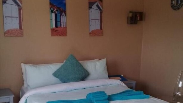 Un lit bien fait, avec une literie blanche et des serviettes bleues, une petite table de chevet et des œuvres d’art murales représentant des maisons.