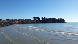 Una playa costera con olas suaves lamiendo la orilla, un rompeolas rocoso y una hilera de edificios al fondo.