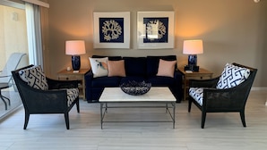 Un salón con un sofá azul oscuro, dos sillones, una mesa de centro y dos mesitas de noche con lámparas. Dos obras de arte enmarcadas decoran la pared sobre el sofá.