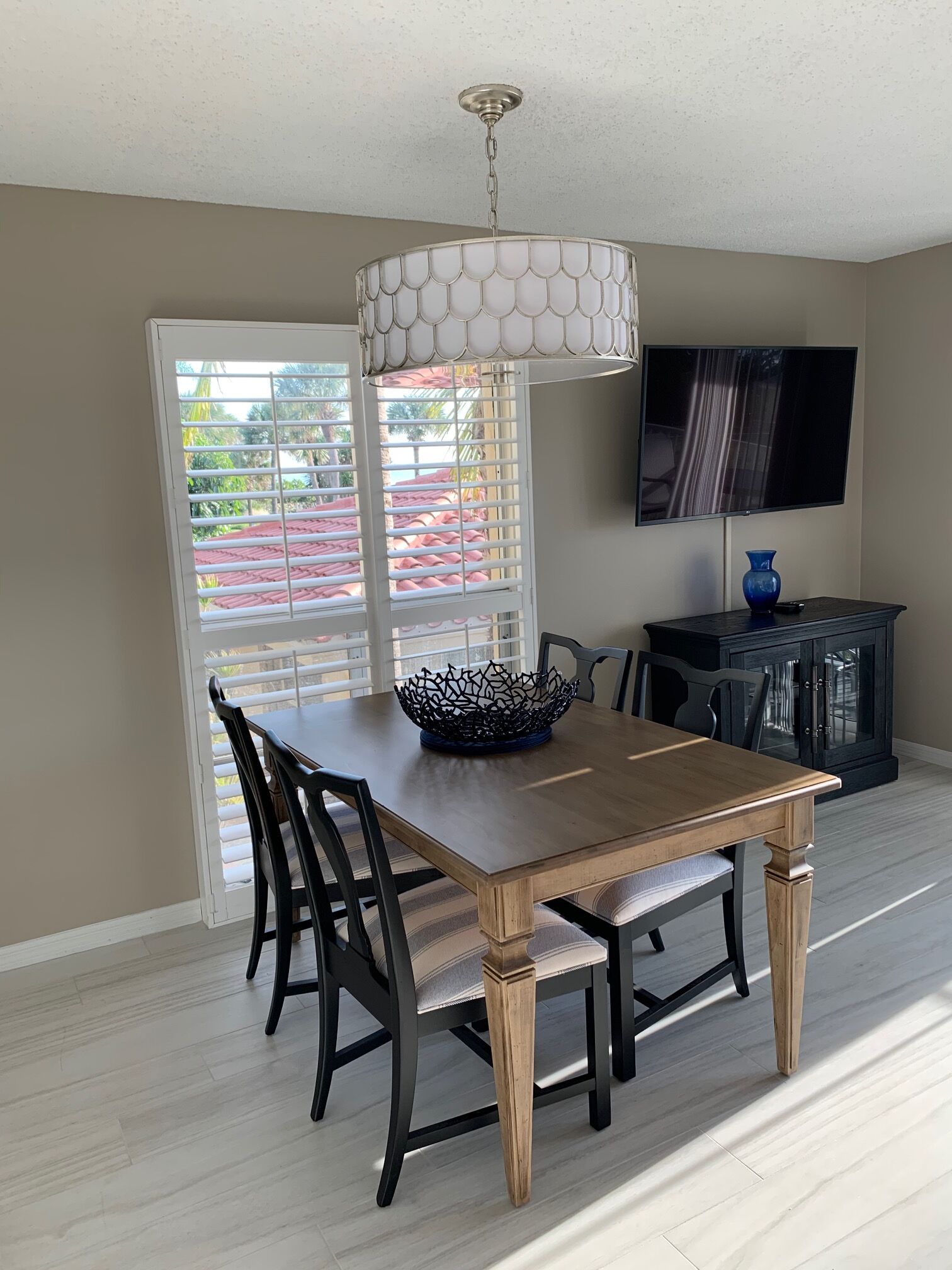 A dining area with a wooden table, black chairs, a decorative bowl, a television mounted on the wall, and a window with blinds.