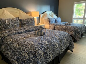 Une chambre avec deux lits, chacun agrémenté d’une parure de lit à motifs floraux bleus assortis et de coussins blancs. On y trouve une table de chevet avec une lampe, ainsi qu’une fenêtre avec des stores.