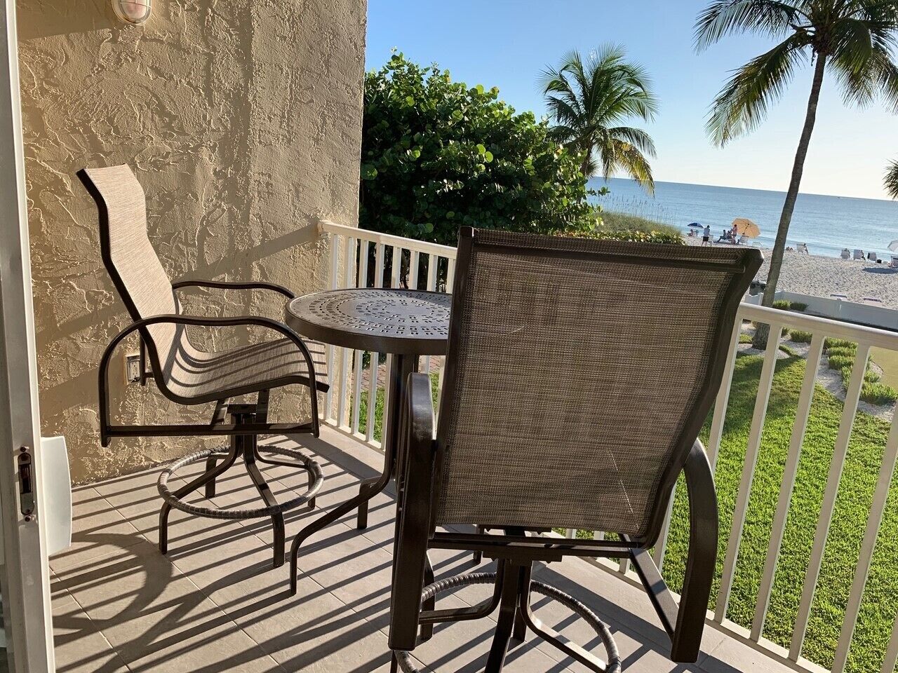 A balcony with a table and two chairs, overlooking a beach with palm trees and people in the distance.