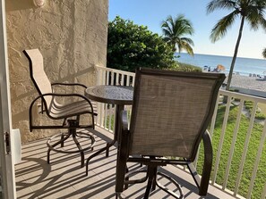 Un balcon avec une table et deux chaises, donnant sur une plage bordée de palmiers et où l’on aperçoit des personnes au loin.