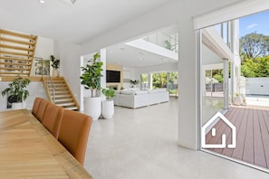Un interior moderno con una mesa de comedor de madera, una escalera y una sala de estar con sofá y ventanas amplias.