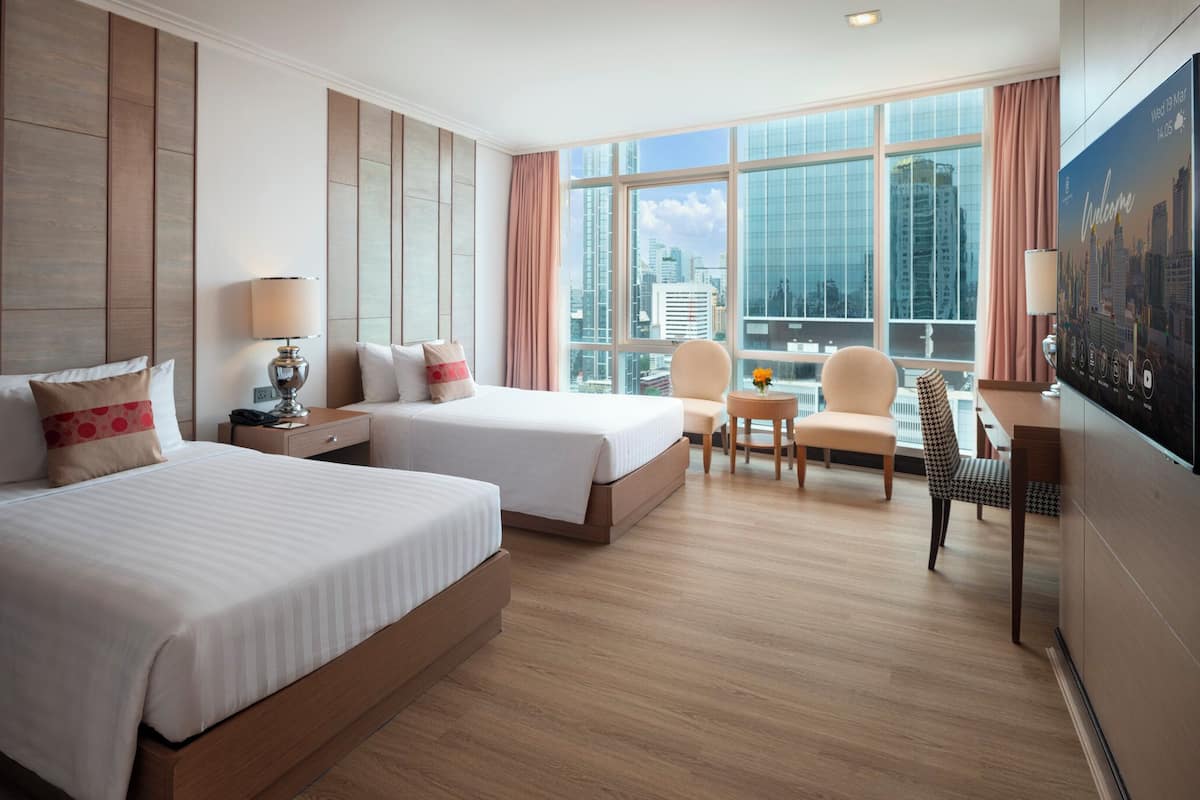 A hotel room with two beds, a desk, a chair, and a large window offering a city view.