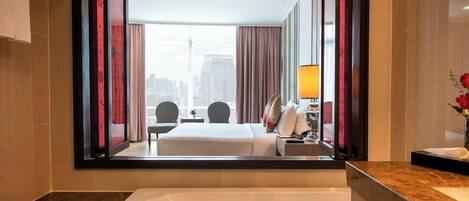 Une chambre d’hôtel dotée d’une grande fenêtre, d’une baignoire, d’une coiffeuse avec un miroir et d’un lit offrant une vue sur le paysage urbain.