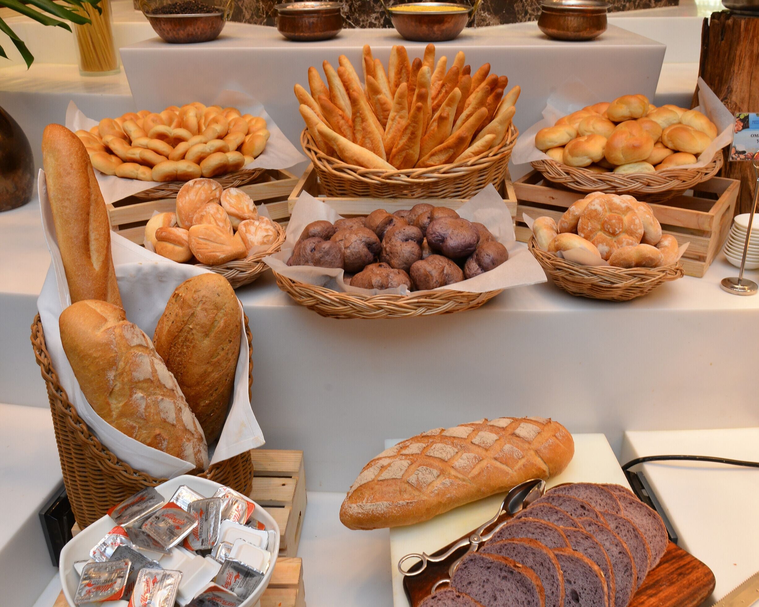 A table with various types of bread and pastries, including baguettes, croissants, and rolls, arranged in baskets and on a cutting board.