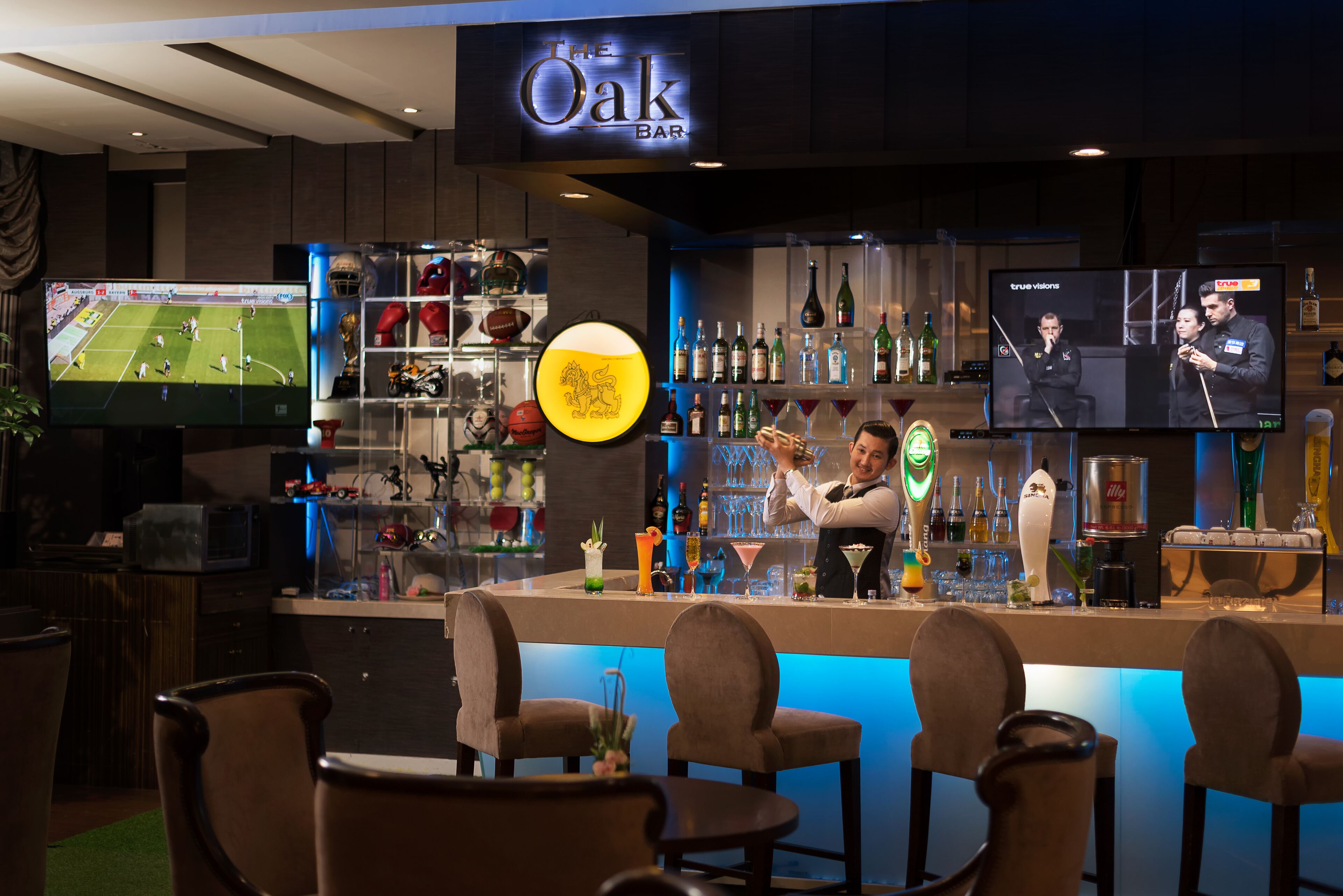 Zona de bar con una televisión mostrando un evento deportivo, barra con un cantinero y diferentes botellas y vasos.