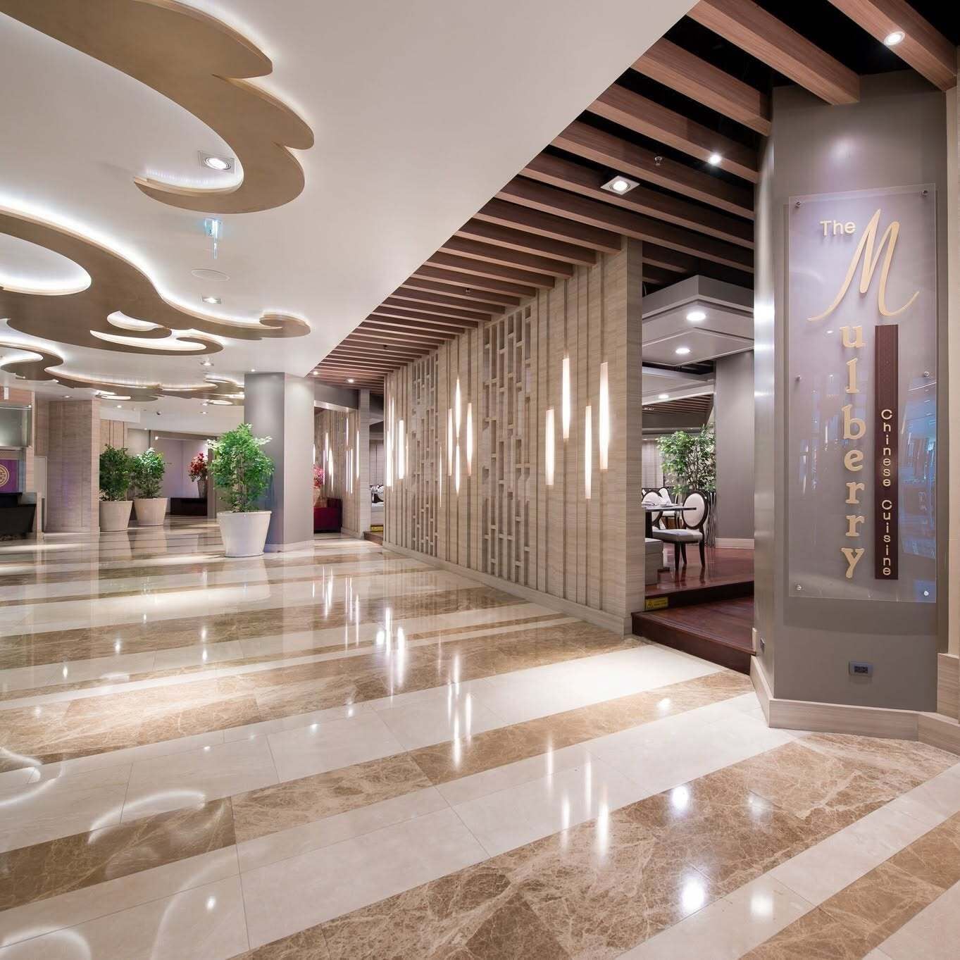 A modern hotel lobby with a marble floor, a reception desk, and a wall with a sign reading 'The Mulleberry Chinese Cuisine.'