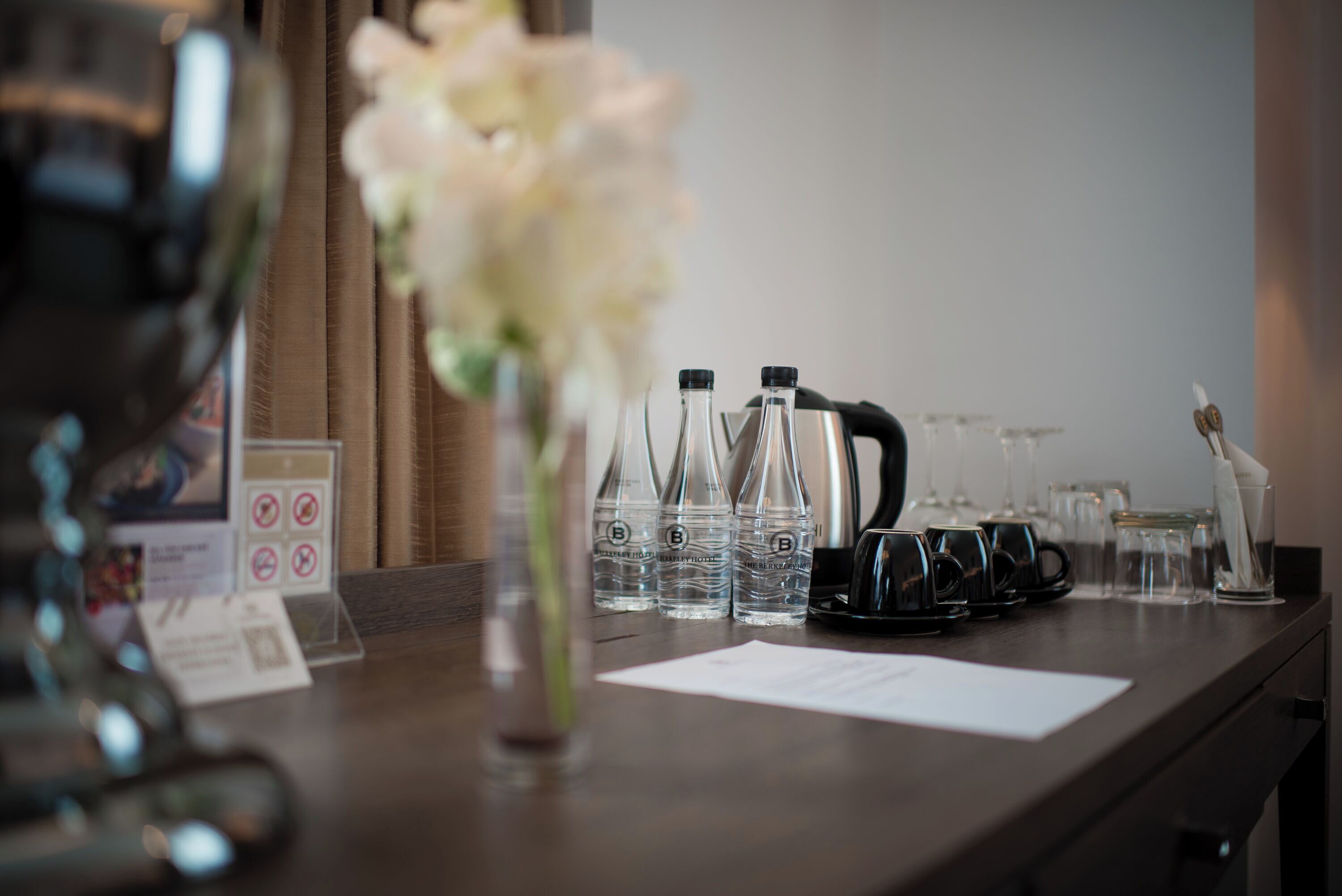 Zona de recepción de un hotel con un mostrador de madera, un jarrón con flores blancas y varias botellas y vasos de vidrio.
