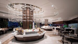 Een moderne hotellobby met een centraal ontbijtbuffet, zitgedeeltes en decoratieve verlichting.