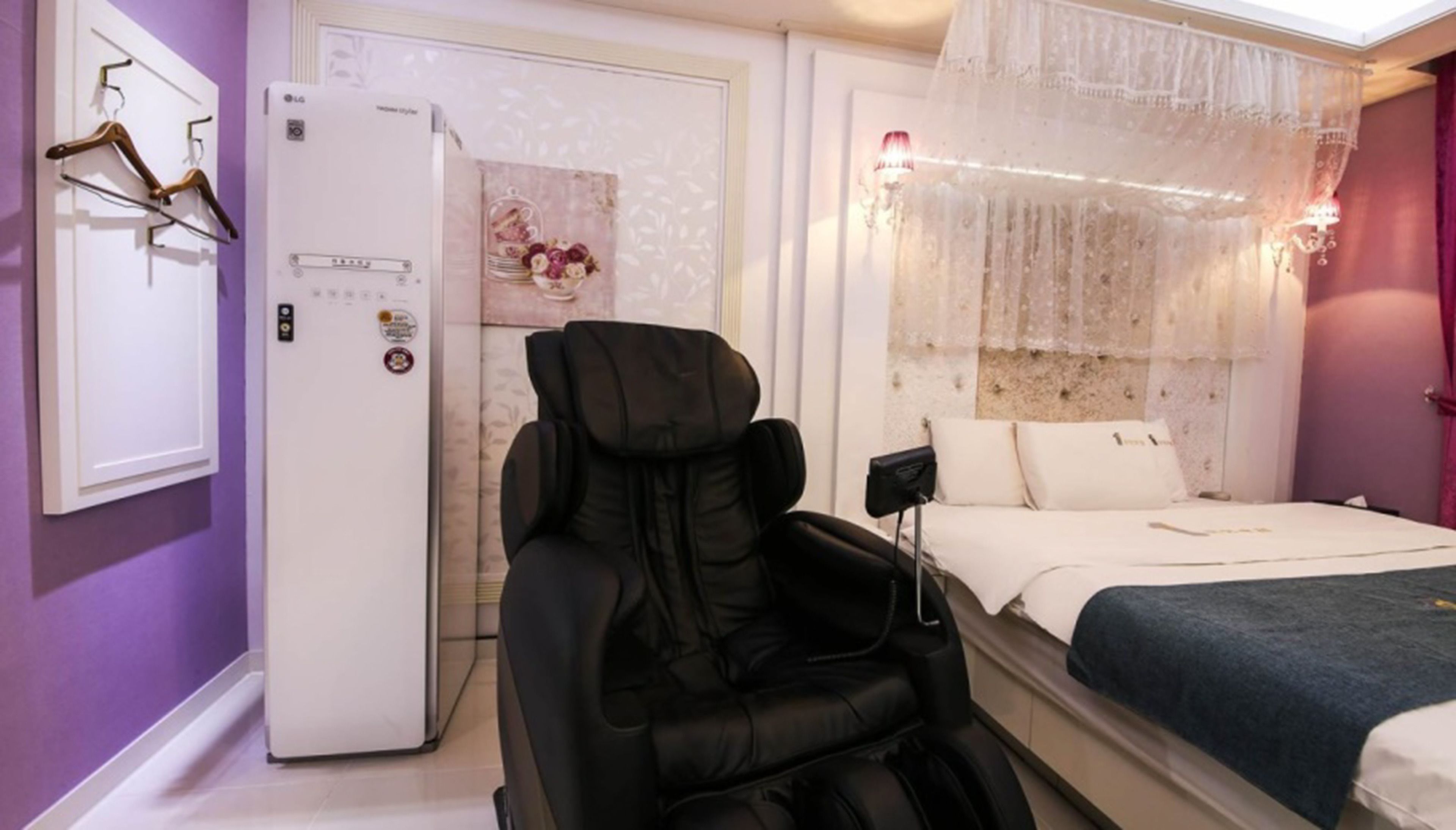 A bedroom with a bed, a massage chair, a refrigerator, and a window with sheer curtains.
