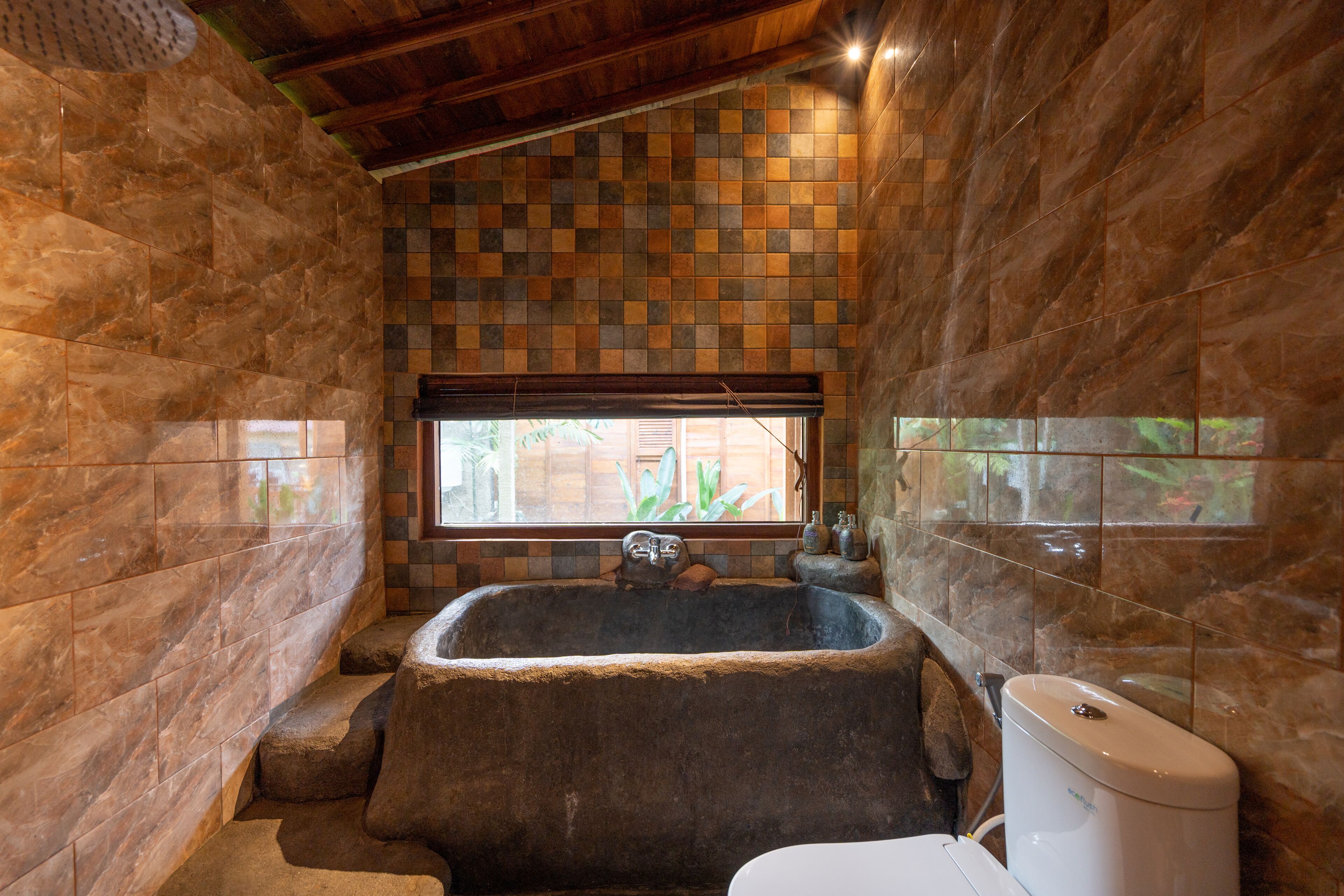 A bathroom with a large stone bathtub, a window with a view of greenery, and a tiled wall with a checkered pattern.