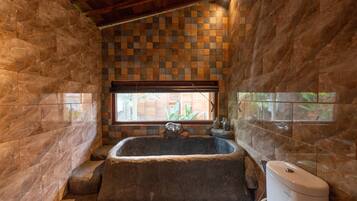 A bathroom with a large stone bathtub, a window with a view of greenery, and a tiled wall with a checkered pattern.