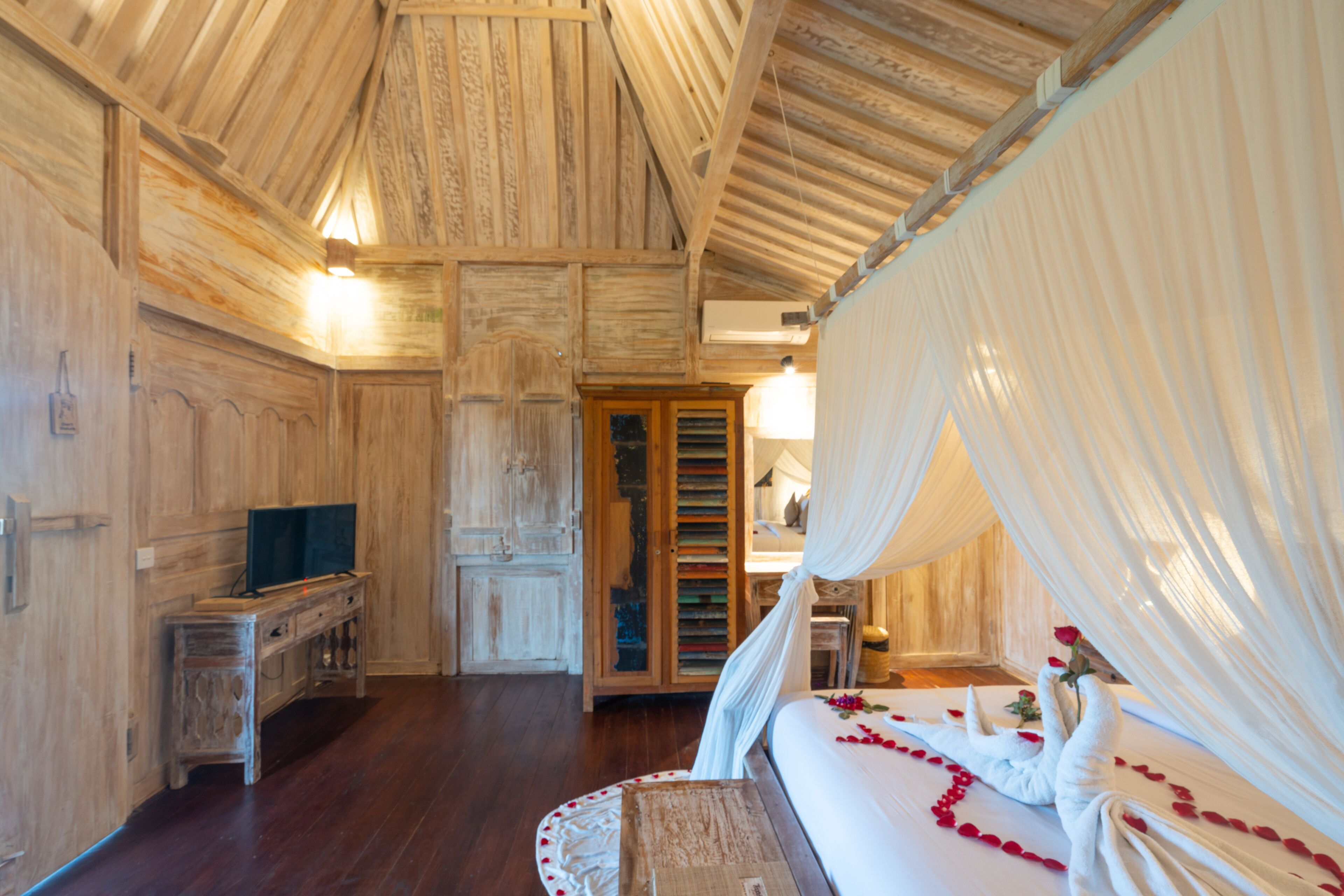 Une pièce avec un plafond en bois, un lit recouvert de draps blancs et parsemé de pétales de rose, une armoire en bois et un téléviseur posé sur un meuble en bois.