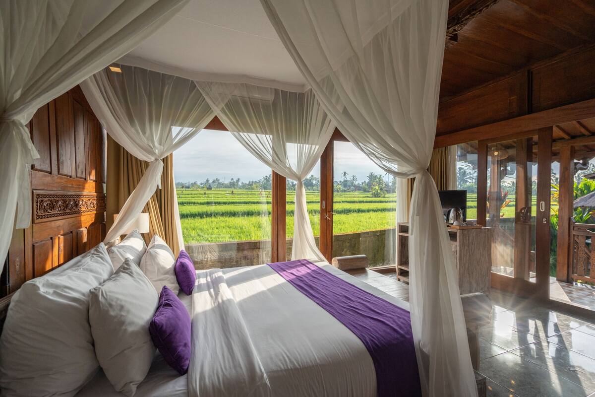 A bedroom with a large bed, white and purple bedding, and a view of green fields through sheer curtains.