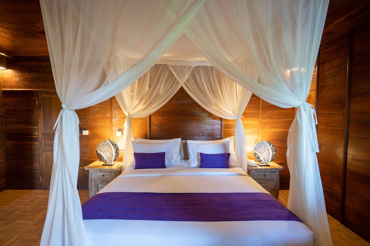 A four-poster canopy bed with white linens and purple accents, wooden headboard, bedside lamps, and a wooden floor.