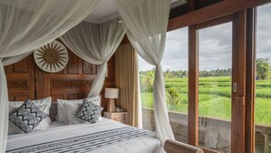 A bedroom with a canopy bed, a wooden bench, and a view of a green field through a glass door. - Uma Sebatu Villas Ubud (Sebatu)