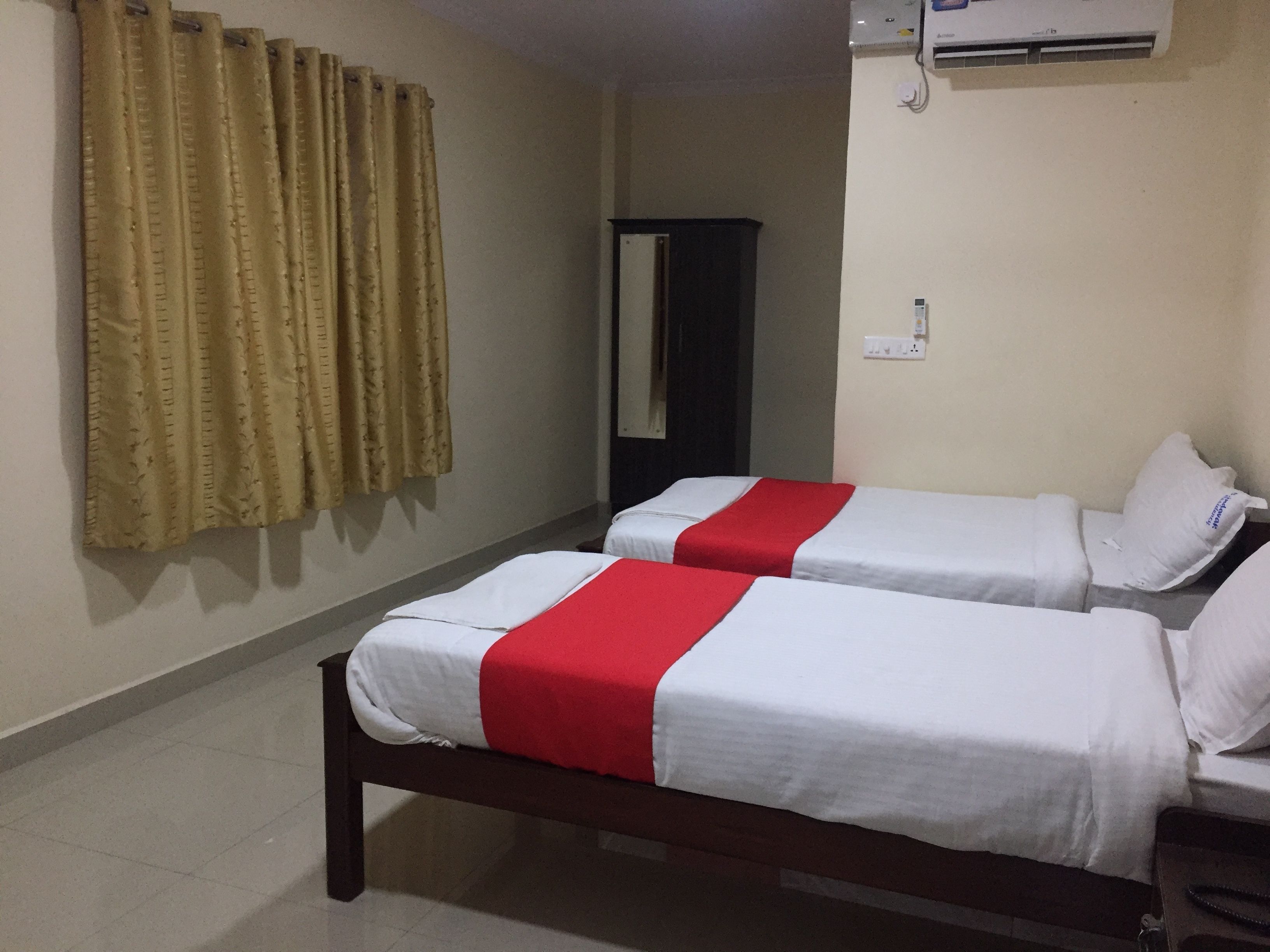 Habitación de hotel con dos camas individuales, cortinas color beige, un armario de madera oscura y un equipo de aire acondicionado en la pared.