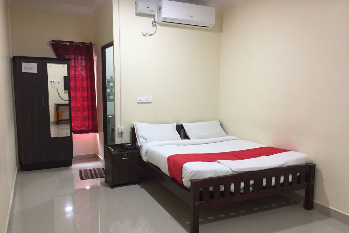 A bedroom with a bed, a nightstand, a wardrobe, a mirror, and an air conditioning unit.