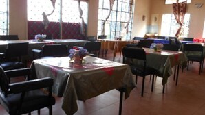 A dining area with multiple tables and chairs, large windows with curtains, and a tiled floor.
