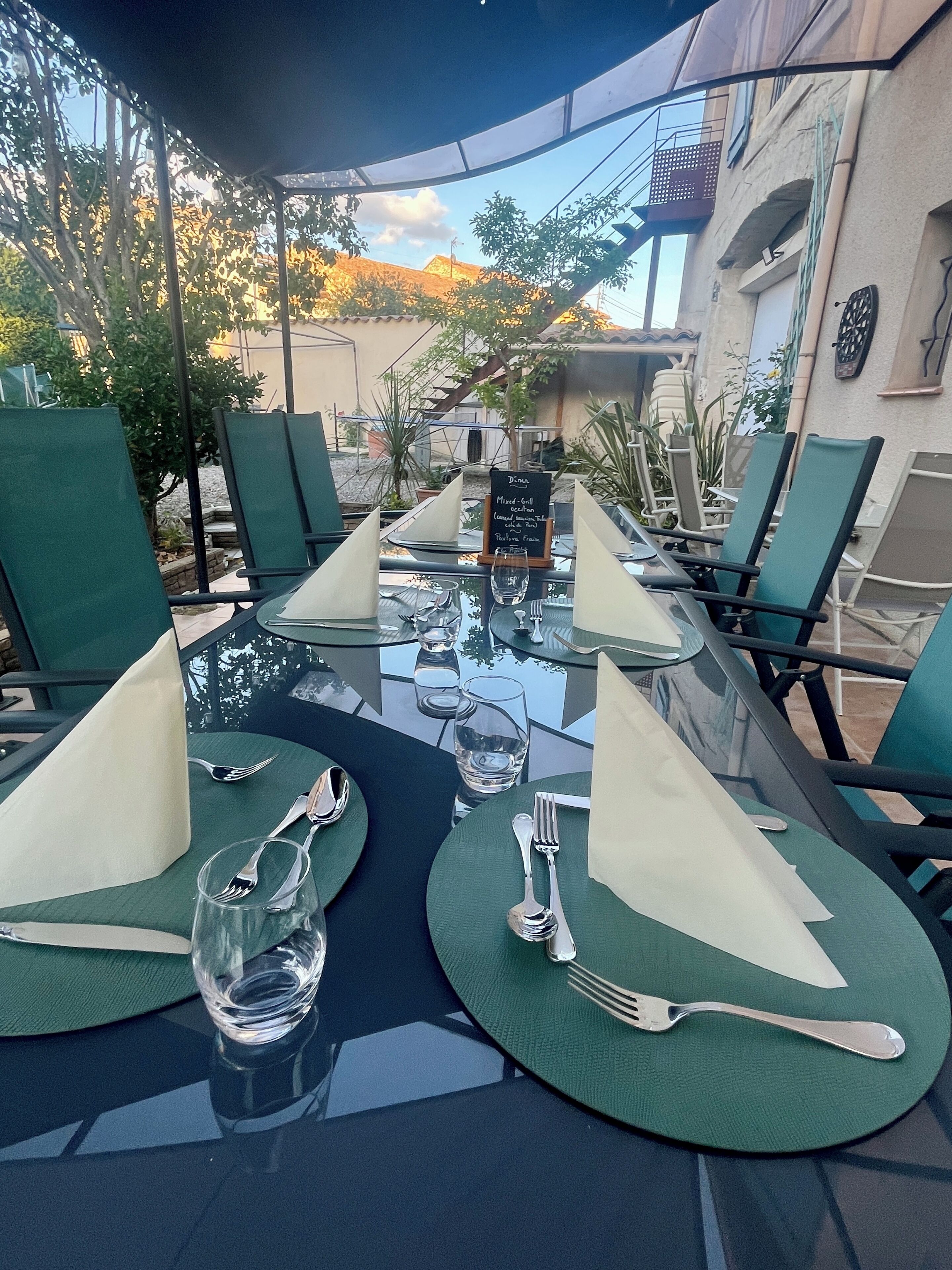 Une table dressée pour un repas en plein air, avec des chaises vertes, une table en verre et un panneau indiquant le menu.