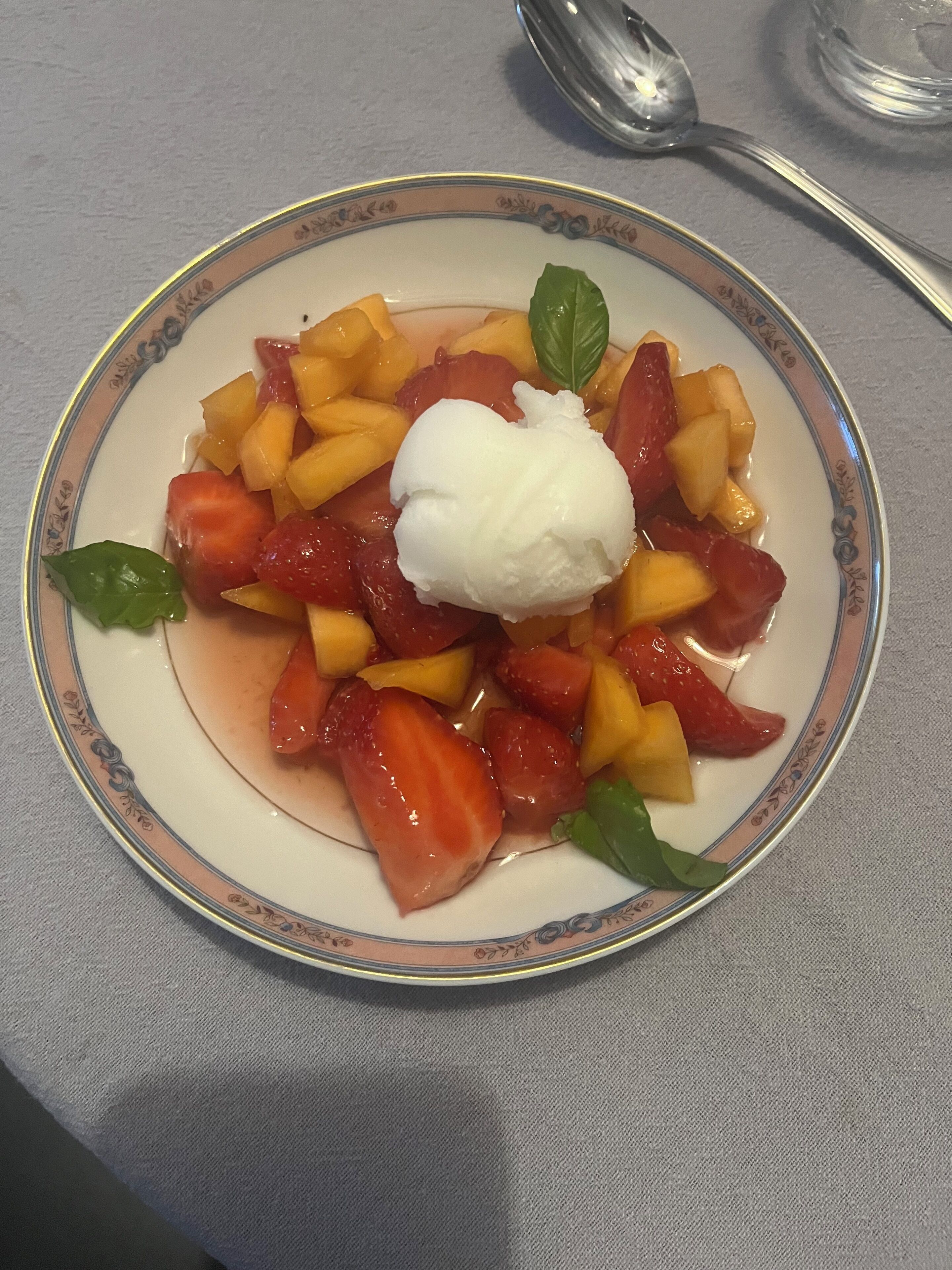 Uma salada de frutas com morangos, mangas e uma bola de sorvete, decorada com uma folha de manjericão.