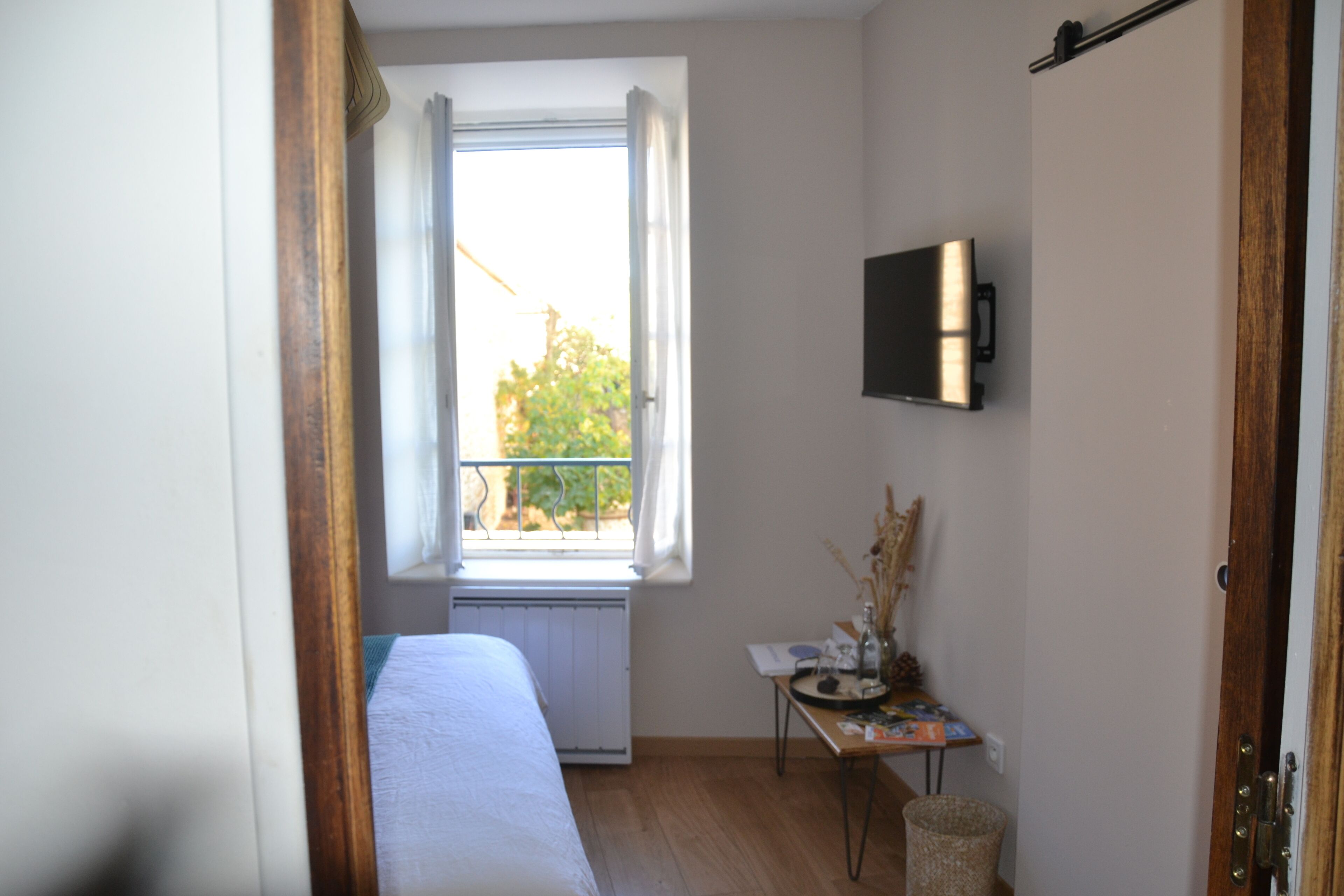 Een kamer met een bed, een klein tafeltje met spullen, een radiator en een raam met uitzicht op groen.