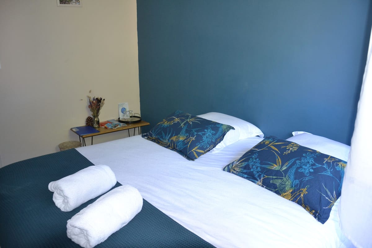 A neatly made bed with blue floral pillows and white linens, a small table with a vase and a book, and a plain wall.
