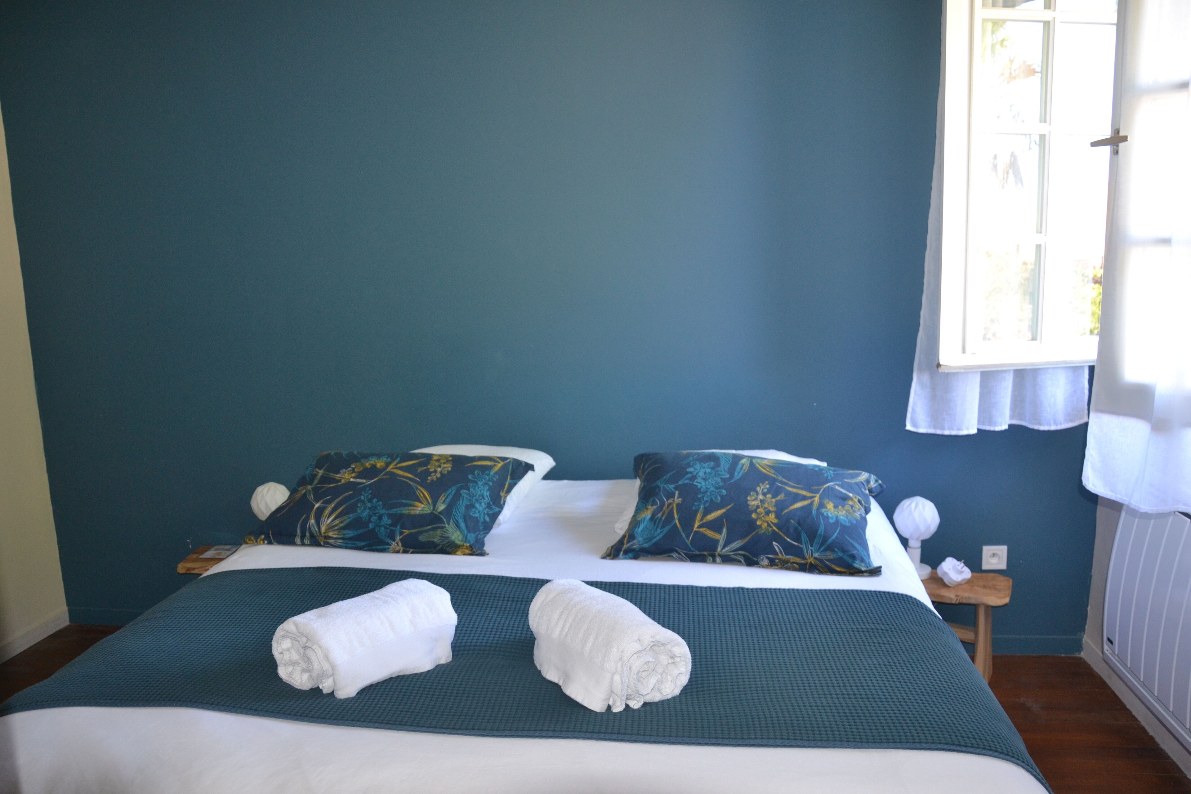 Una cama bien hecha con dos almohadas, una pared azul y una ventana con cortinas.