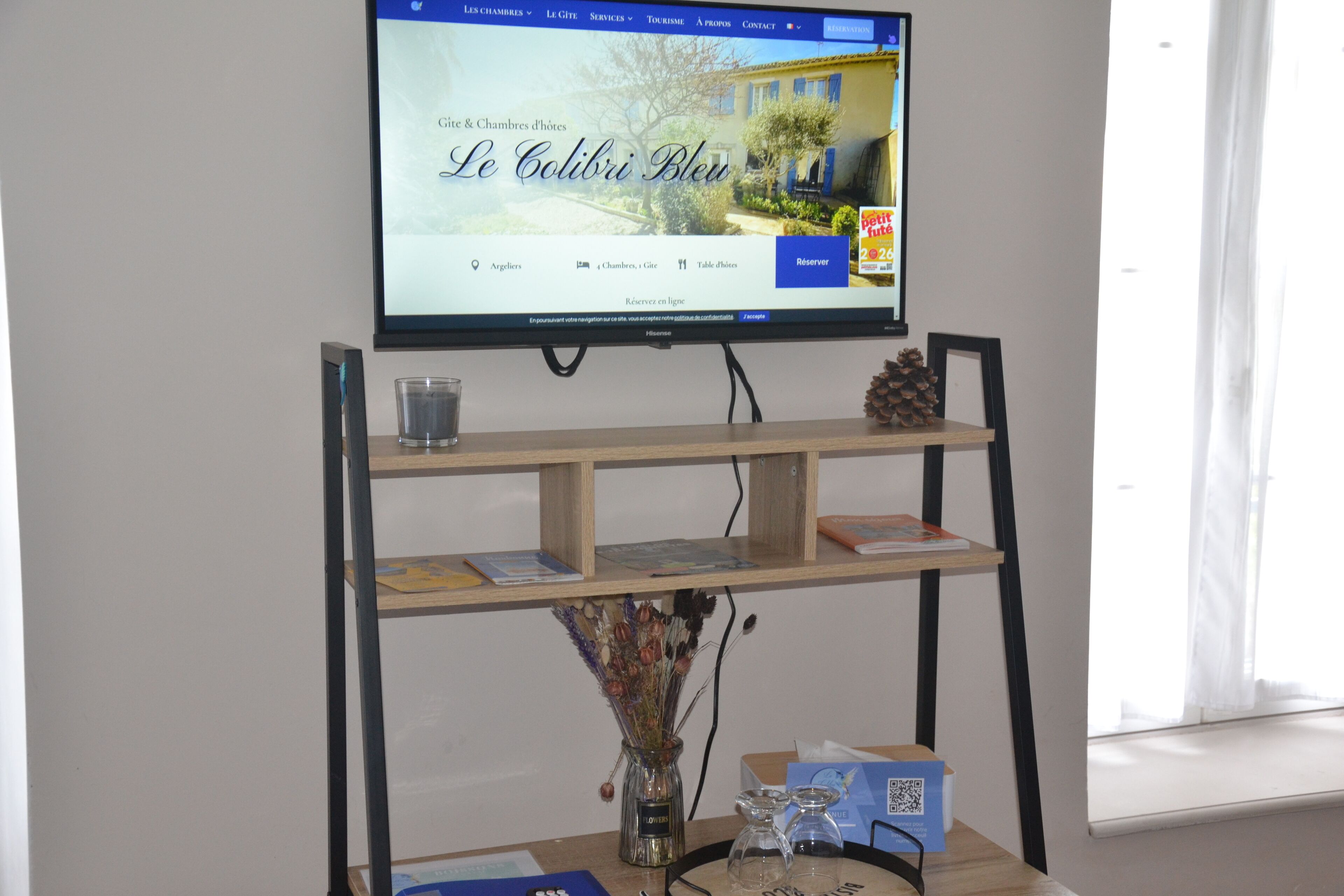Habitación de hotel con un televisor montado en un soporte mostrando una página web. Hay dos estantes debajo del televisor, uno con un vaso y un libro, y otro con un jarrón de flores secas.