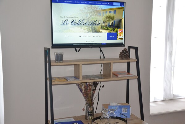 Habitación de hotel con un televisor montado en un soporte mostrando una página web. Hay dos estantes debajo del televisor, uno con un vaso y un libro, y otro con un jarrón de flores secas.