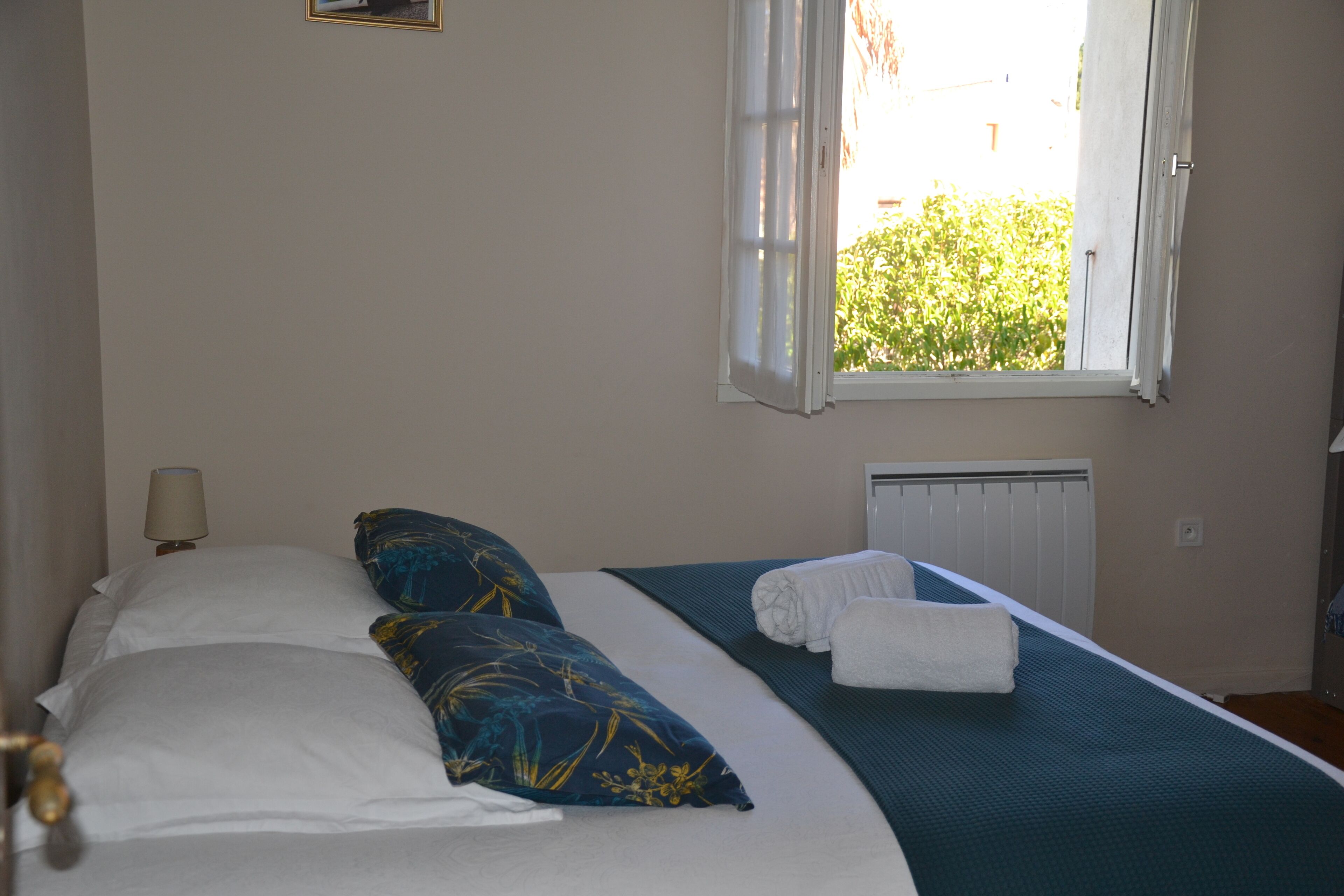 A neatly made bed with white and blue pillows, a teal bedspread, and a white towel on the side. There is a window with white curtains, a wall-mounted lamp, and a radiator.