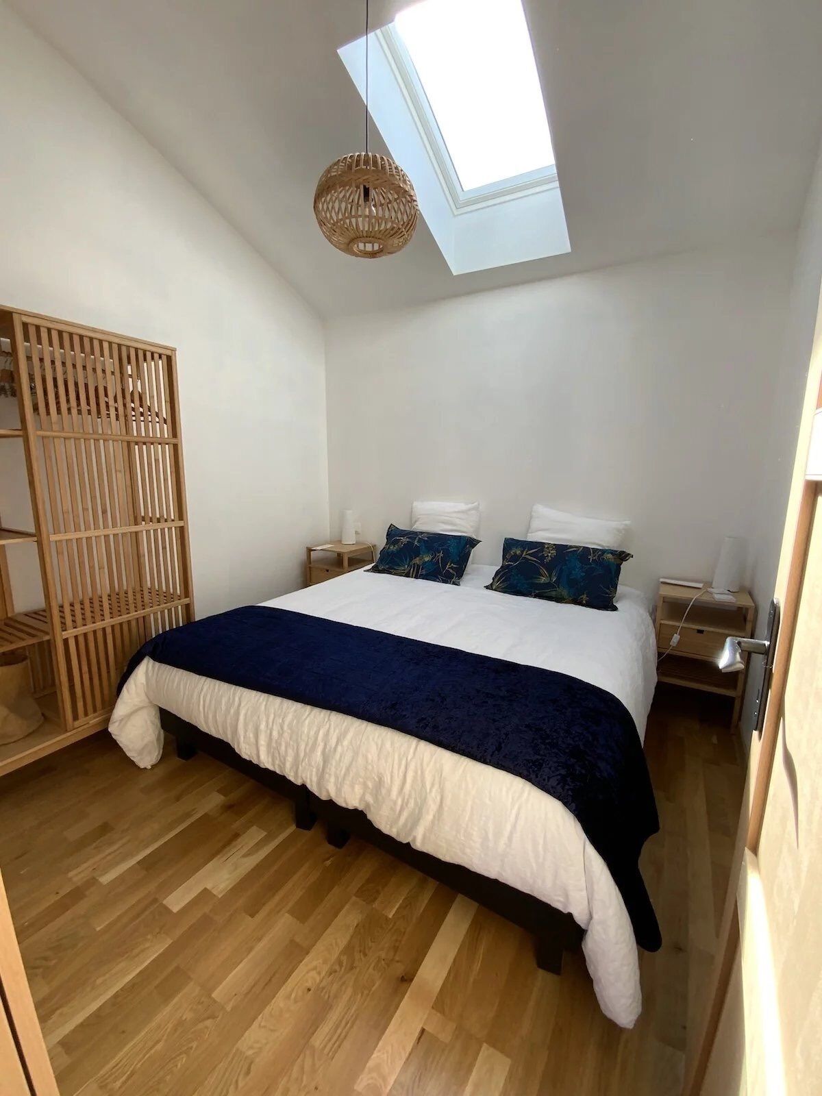 A bedroom with a skylight, a bed with a white and navy blue bedspread, wooden flooring, and a wooden partition with vertical slats.