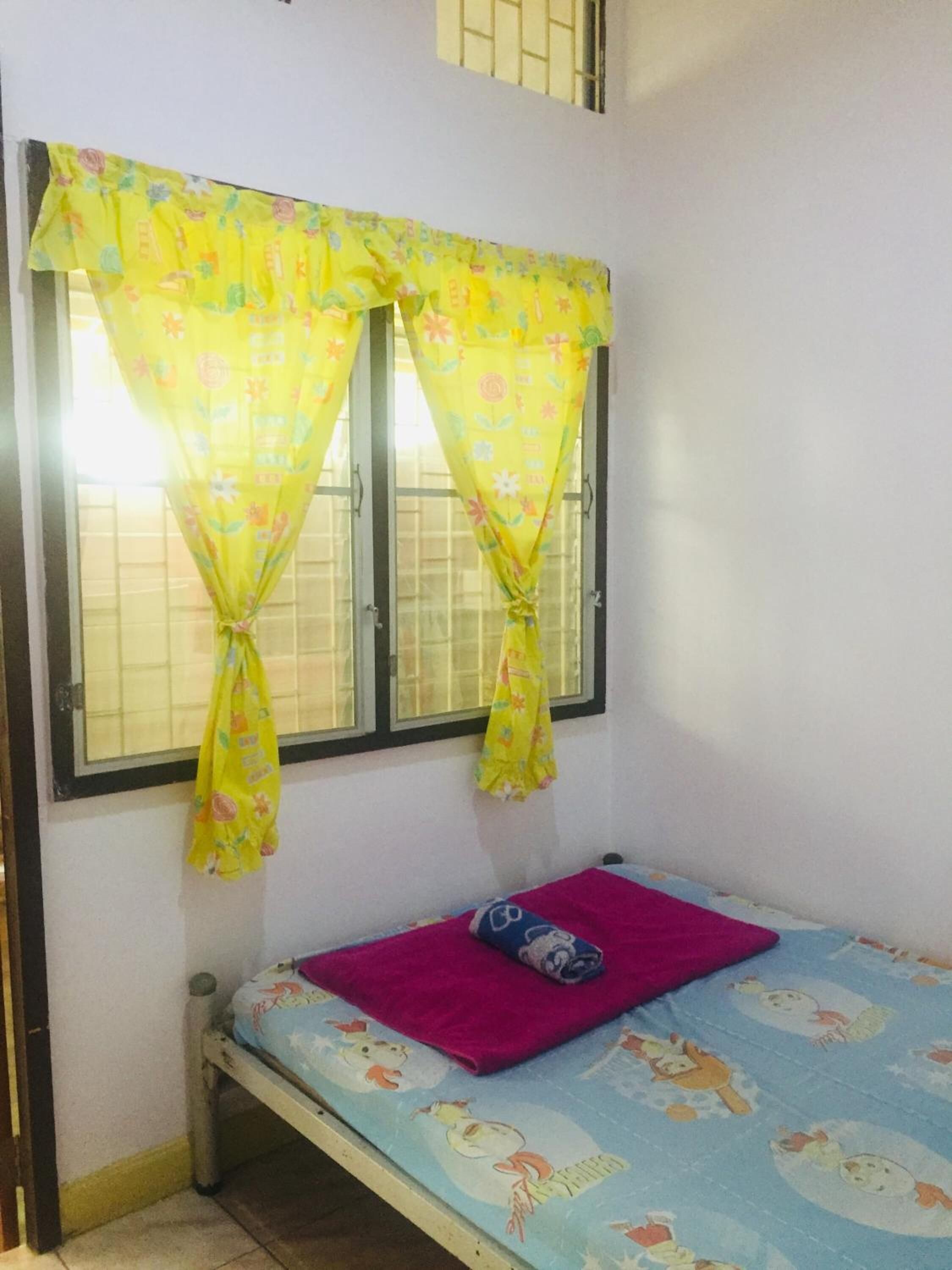 Quarto pequeno com uma cama de solteiro, janela com cortinas amarelas e um cobertor rosa na cama.