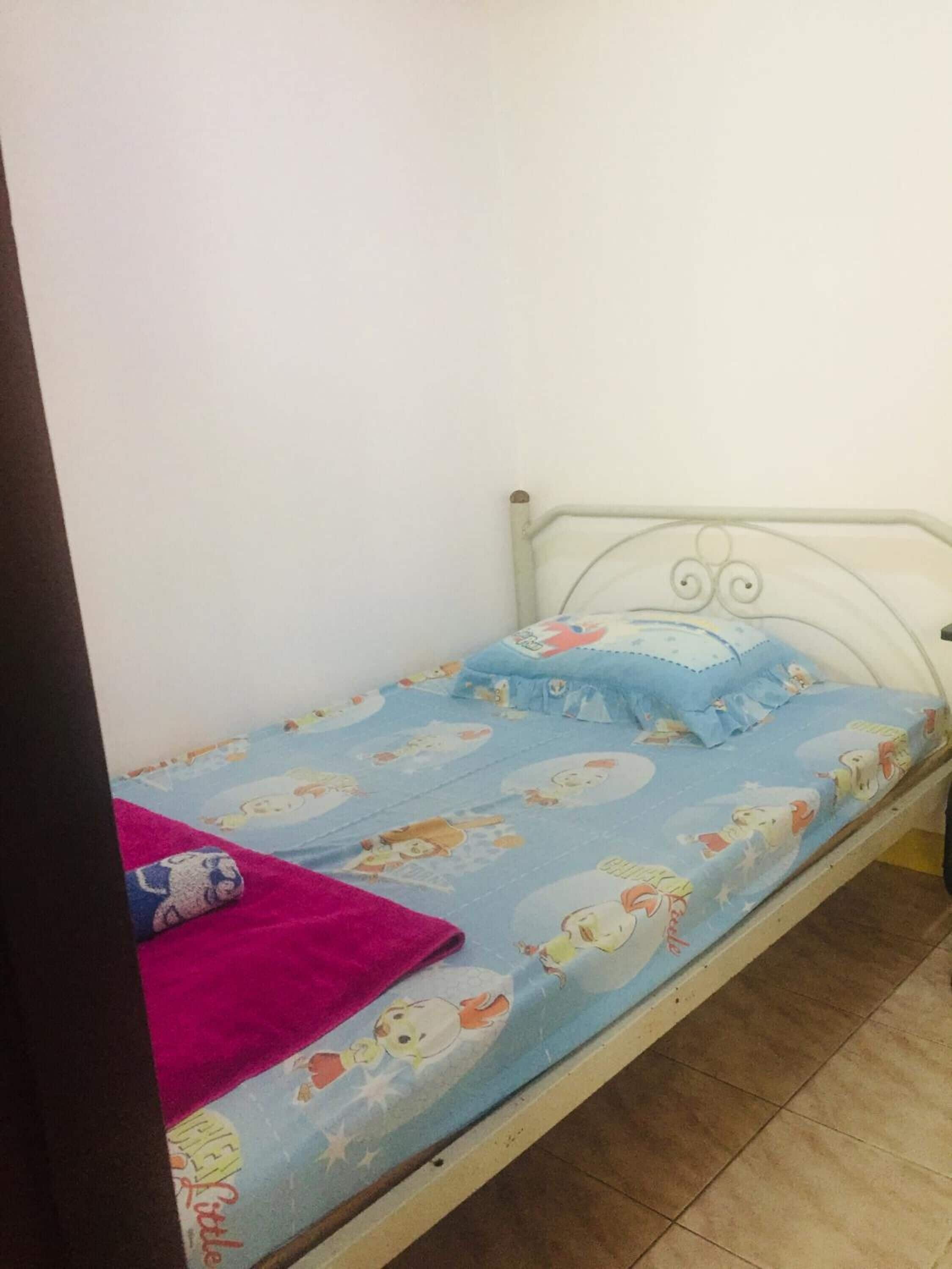 Una cama individual con estructura metálica blanca, colchón azul con personajes de dibujos animados y una manta rosa.