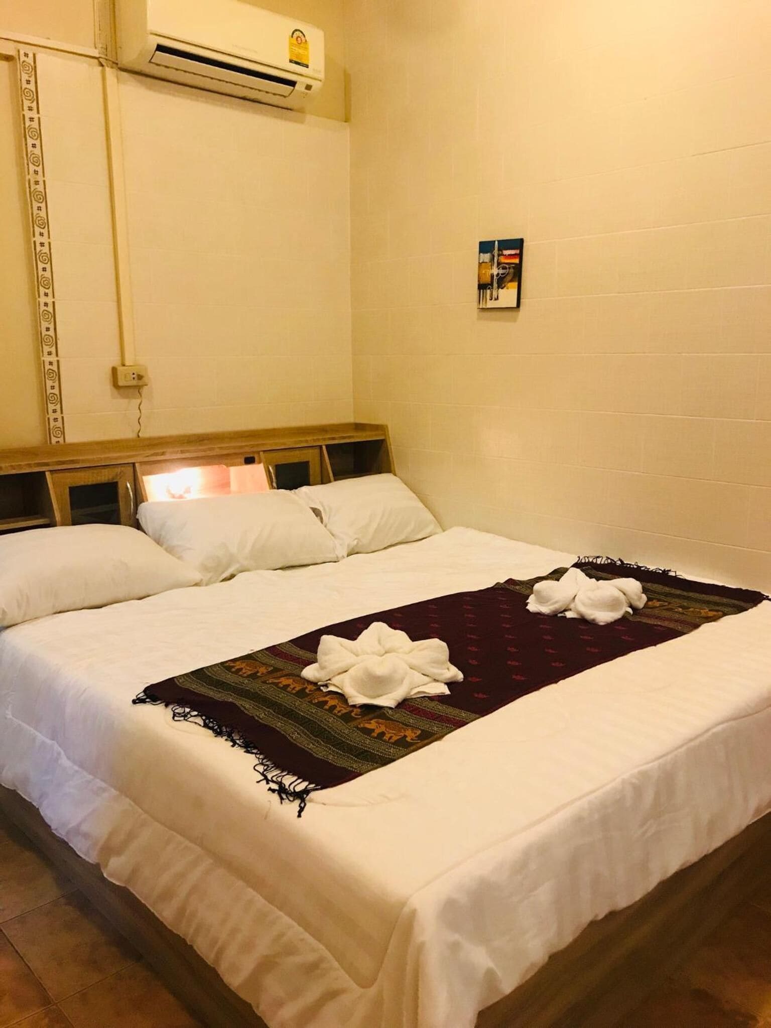 A neatly made bed with white linens and a dark patterned blanket, a wooden headboard, and a wall-mounted air conditioner.