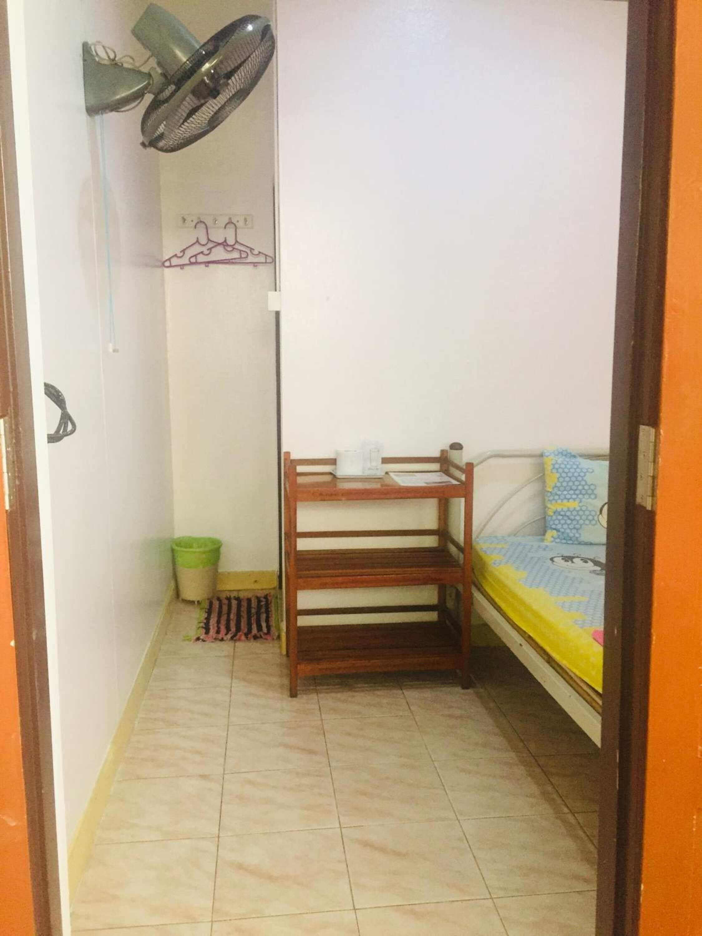 A small, compact room with a bed, a wooden bench, a green trash bin, and a wall-mounted fan.