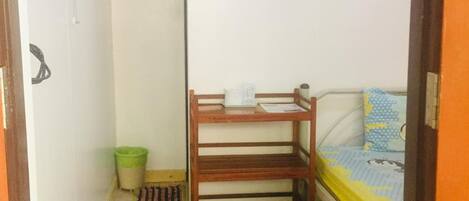 A small, compact room with a bed, a wooden bench, a green trash bin, and a wall-mounted fan.