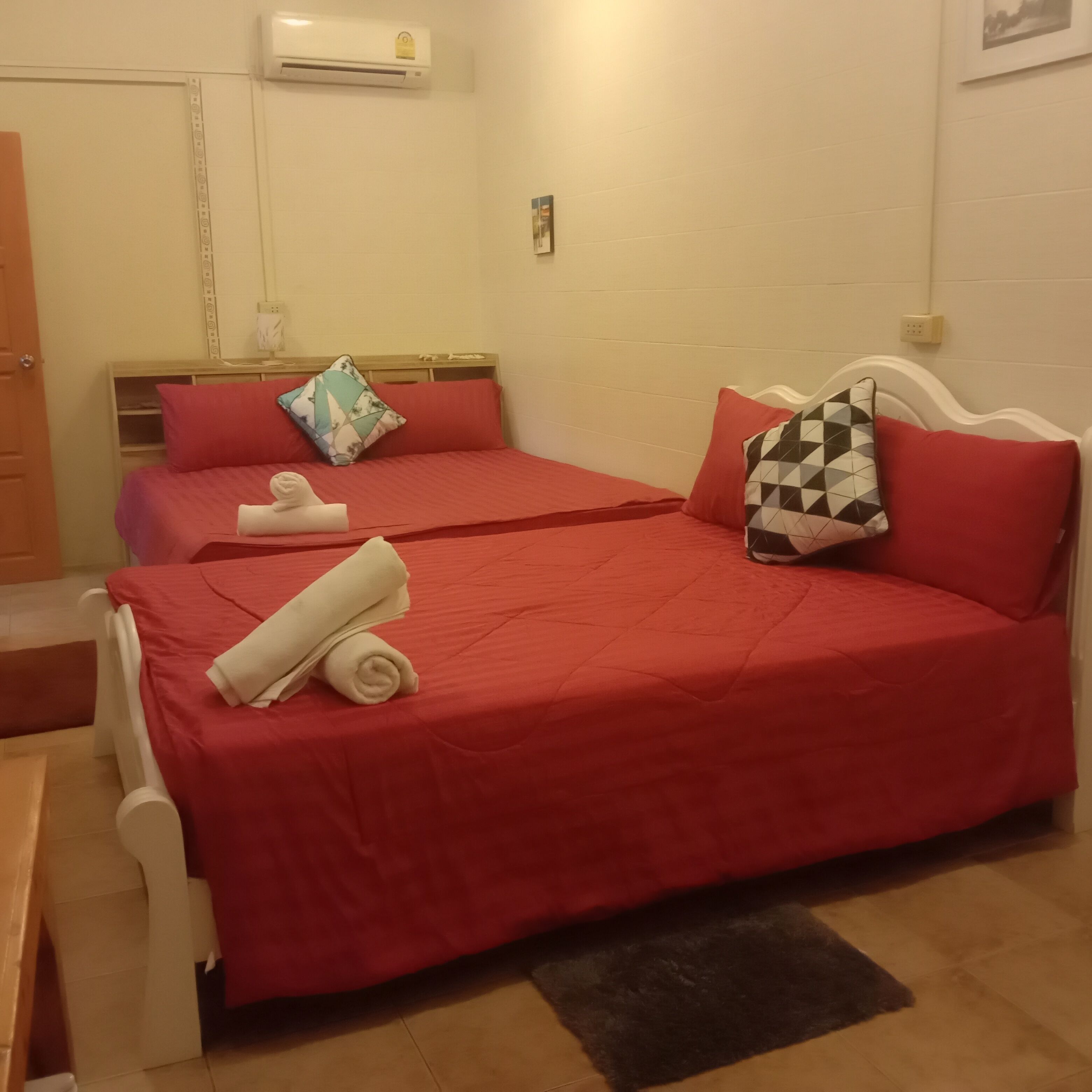 A hotel room with two beds, each with red bedding and pillows, a white headboard, a small bedside table, and a wall-mounted air conditioner.