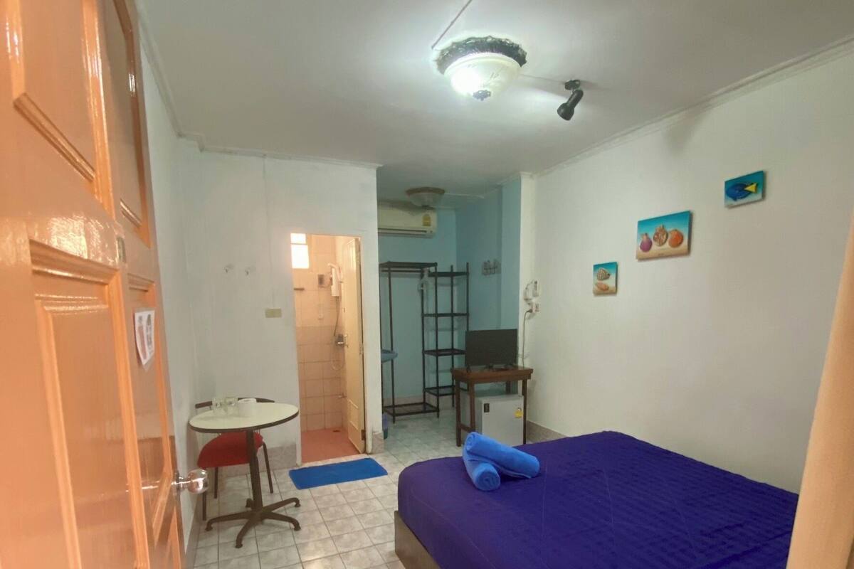 Ein kleiner, sauberer Raum mit einem Bett, einem Schreibtisch und einem Stuhl. Eine Tür ist links, ein Fenster rechts.