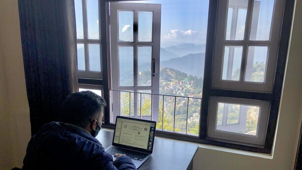 Uma pessoa sentada à mesa com um laptop, de frente para uma janela com vista de uma paisagem montanhosa.