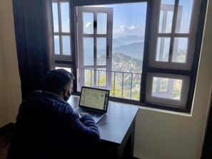 A person sitting at a desk with a laptop, facing a window with a view of a mountainous landscape. - The Griffon's Nest (Gangtok)