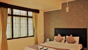 Quarto de hotel com uma cama grande, janela com cortinas, mesa de cabeceira e luminária fixada na parede.