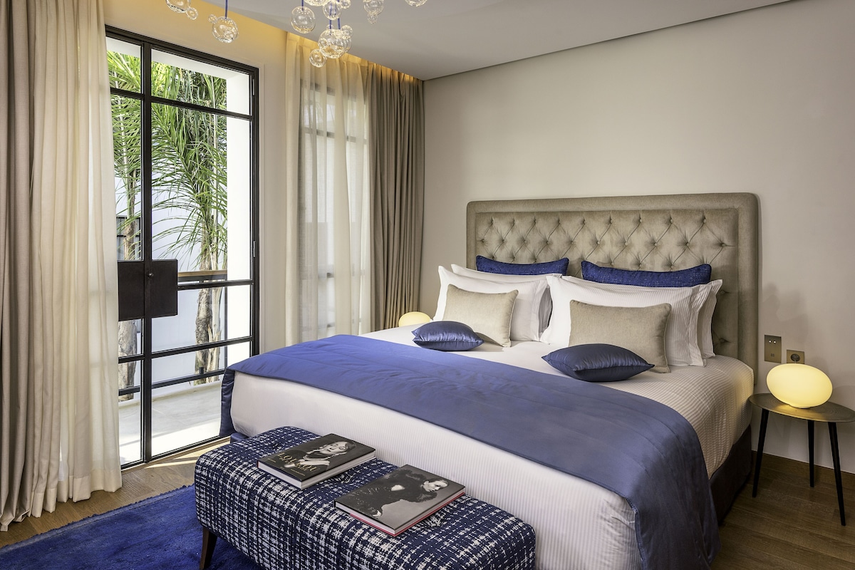 A bedroom with a large bed, a bedside table with a lamp, and a view of palm trees through a window.