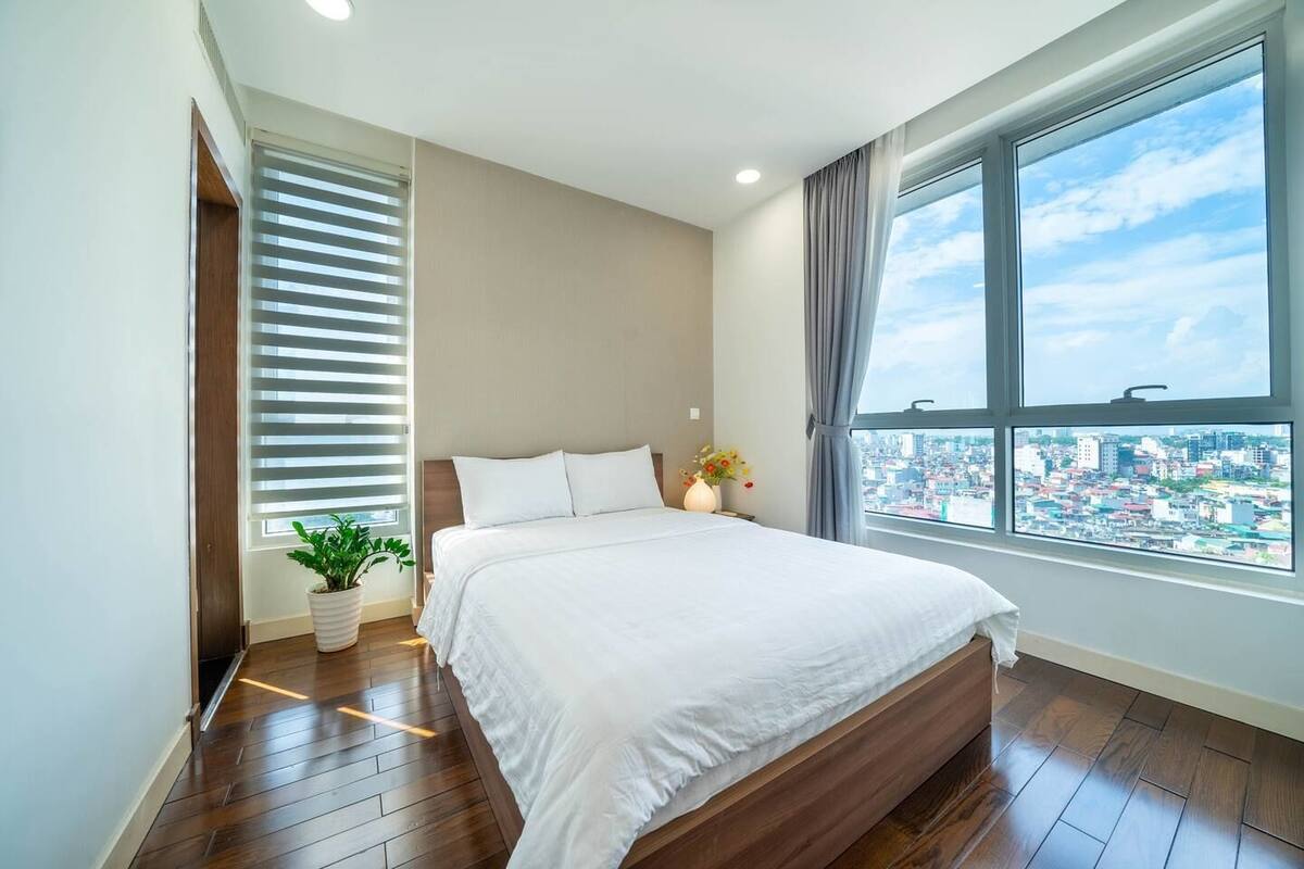 Une chambre moderne avec un grand lit, une plante en pot et une vue sur la ville par la fenêtre.