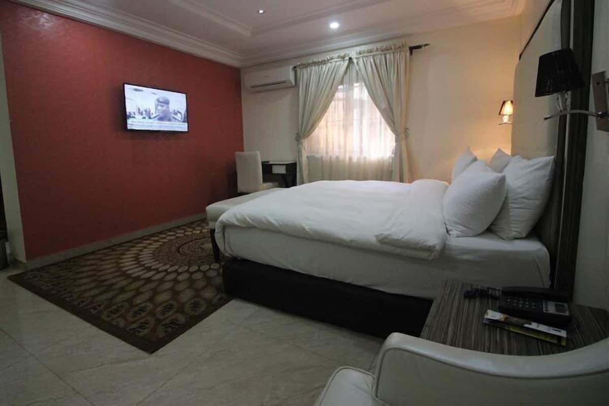 A hotel room with a bed, a television mounted on the wall, a desk with a chair, and a window with curtains.