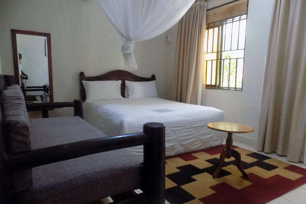 Quarto de hotel com cama, mesinha redonda, cadeira, espelho e janela com cortinas.